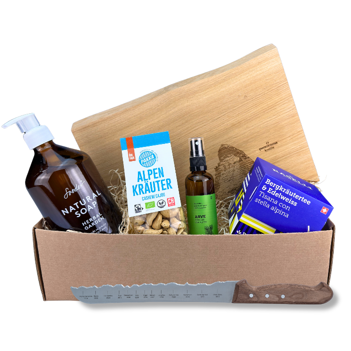 Alpen Bergwelt Premium Geschenkbox "Matterhorn" befüllt mit Soeder Natural Soap Herbal Garden Seife, Pakka Alpenkräuter Cashews, Arve Raumduft und kissenspray, BIO Raselli Bergkräuter und Edelweiss Tee, rustikales Matterhorn Schneidebrett von PanoramaKnife und einem Brotmesser. Vor weissem Hintergrund.
