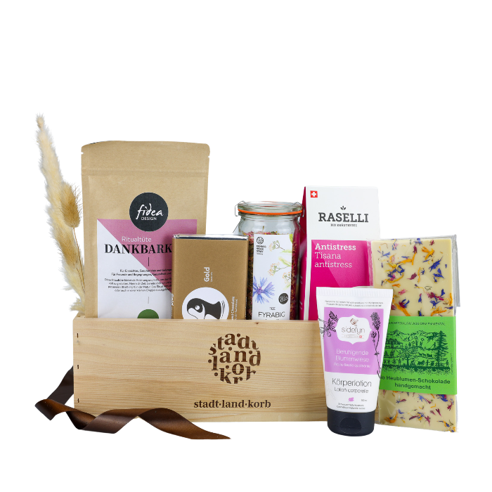 Fidea Ritualtüte Dankbarkeit, Taucherli Gold Schokolade, Fyrabig Tee im Weckglas, Bio Raselli Antistress Tee, Heublumen Schokolade un d Sidefyn Körperlotion in einem Stadt Land Korb Geschenkkorb aus Holz vor weissem Hintergrund.