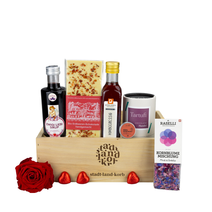 Ewigi Liebi Geschenkkorb mit Ewigi Liebi Sirup, Bio Erdbeeren Schokolade, Himbeeressig, Bärgmüntschi Lipgloss, Mandel Tartufi und Bio Rasellli Kornblumen Mischung in einem Stadt Land Korb Geschenkkorb aus Holz. Eine Rose und Lindt Schokoherzen vordran. Vor weissem Hintergrund.