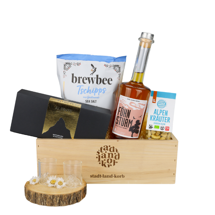 Föhnsturm Geschenkkorb vor weissem Hintergrund befüllt mit Brewbee Sea Salt Tschipps, Alpinte Matterhorn Schnappsgläser, Föhnsturm Whisky und Alpenkräuter Cashews