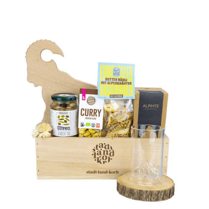 Genuss trifft Stil Geschenkkorb vor weissem Hintergrund befüllt mit Steinbock Apero Brett, Oliven, Curry Cashews, Butter Rädli, Apinte Half Pint matterhorn Glas
