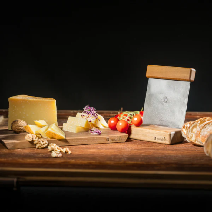Käseschneider Mood Bild mit Käse, Nüssen, Tomaten auf einer Holzplatte und schwarzem Hintergrund.