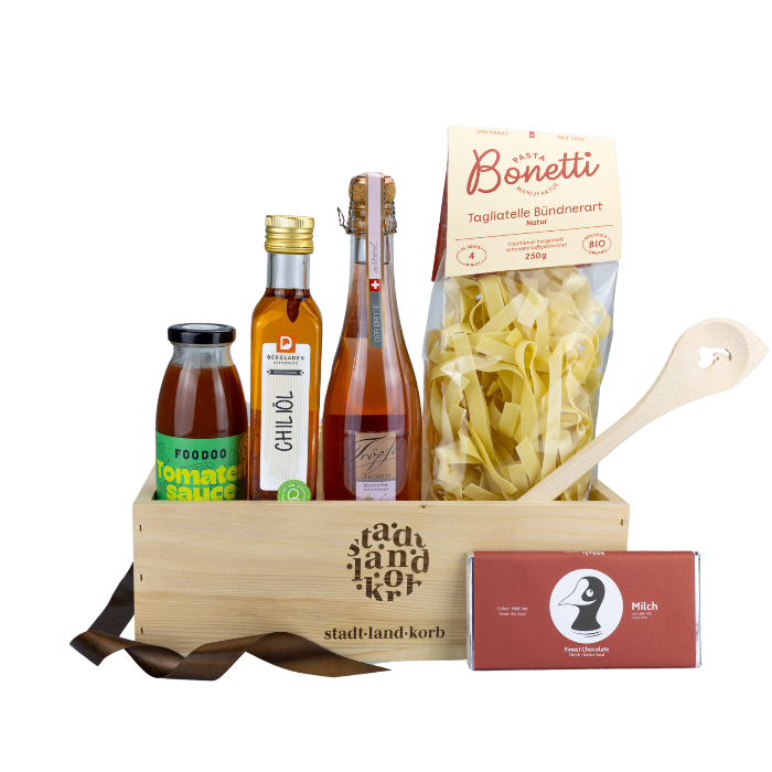 Stadt Land Korb Rosso Geschenkkorb befüllt mit FOODOO Tomatensauce, Chiliöl, Tröpfel der Dritte, Bonetti Tagliatelle, Kochlöffel Steinbock und Taucherli Milchschokolade vor weissem Hintergrund.
