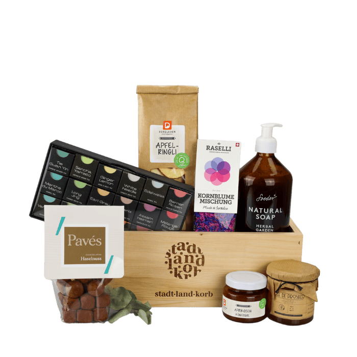Süsse Glücksmomente Geschenkkorb vor weissem Hintergrund befüllt mit Haselnuss Pavés, Geschenkteeschachtel, Apfelringli, Raselli Kornblumen Mischung, Soeder Natural Soap, Konfitüre, Gelée de Prosecco