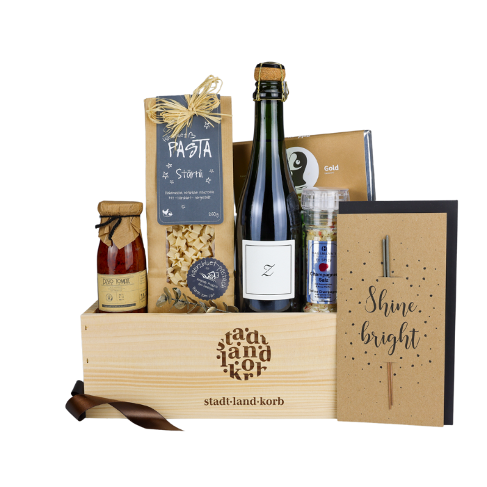 Sparkling Geschenkkorb vor weissem Hintergrund befüllt mit Stärnli Pasta, Tomatenpesto, Zürischum, Champagner Salz, Taucherli Gold Schokolade, Wunderkerzengrusskarte