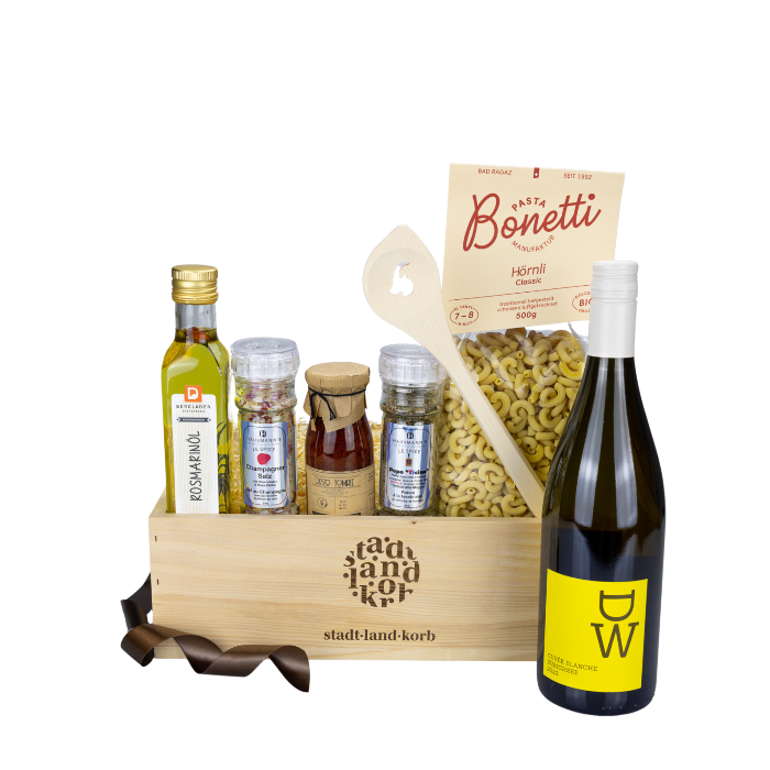 Stadt Land Korb Weisswein Dinner Geschenkkorb befüllt mit Rosmarinöl, Champagner Salz, Tomatenpesto, Pepe Ticino Pfeffer. Bonetti Hörnli, Kochlöffel Steinbock und DW Weisswein vor weissem Hintergrund.
