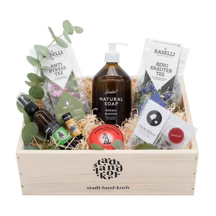 Bergruhe Geschenkkorb: Stadt Land Korb Holzkorb gefüllt mit BIO Raselli Antistress Tee, Bio Raselli Bergkräutertee, Soeder Natural Soap Herbal Garden, Arve Raumduft und Kissenspray, Bärgmüntschi Lipgloss und Badesalz. Vor weissem Hintergrund