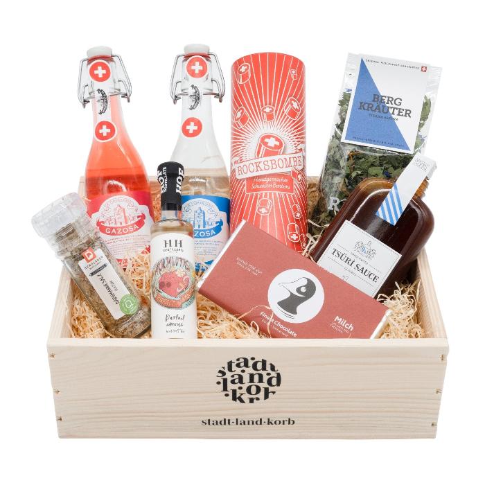 Du bisch eifach Bombe! Geschenkkorb Männerversion: Gazosa Limonaden Duo, Bärgmannesalz, Hofdestillat vom Haldihof aus Luzern, Taucherli Milchschokolade, Tsüri Sauce, Zischmombe Rocks BonBons und Bio Raselli Bergkräutertee in einem Stadt Land Korb Geschenkkorb aus Holz vor weissem Hintergrund.