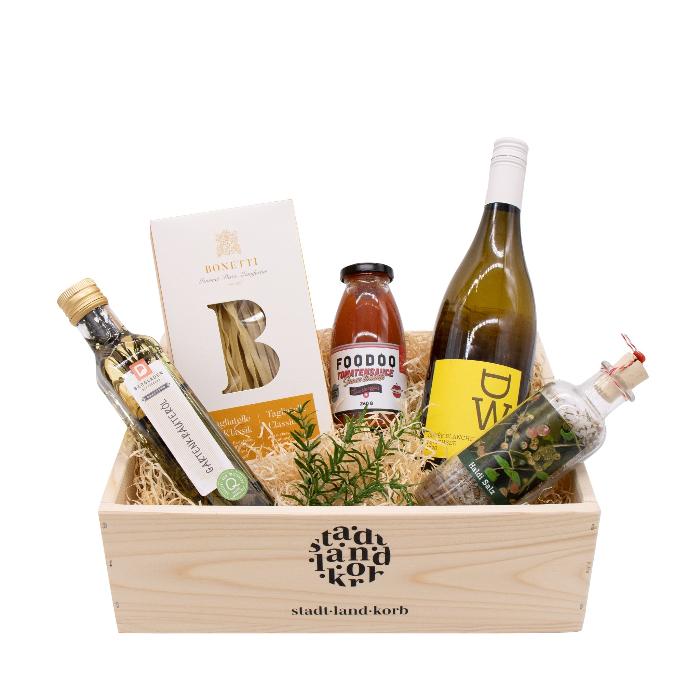 Dinner Blanc Geschenkkorb: Gartenkräuter-Öl, Bonetti Tagliatelle, FOODOO Tomatensauce, DW Weisswein und Haldisalz in einem Stadt Land Korb Geschenkkorb aus Holz vor weissem Hintergrund.