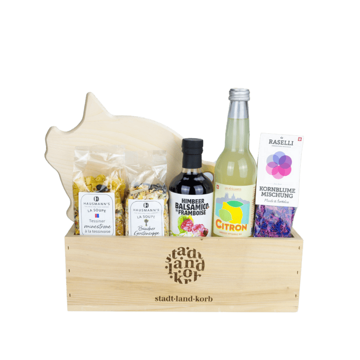 Feinschmecker Geschenkkorb vor weissem Hintergrund befüllt mit Schneidebrett Säuli, Tessiner Minestrone, Bündner Gerstensuppe, Himbeer Balsamico, Zitronen Limonade und essbare Kornblumen