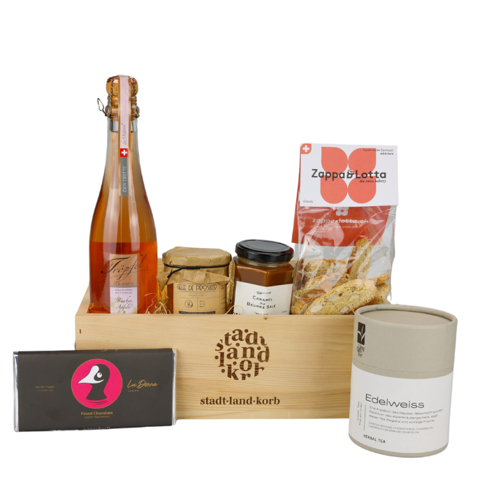 Genuss und Geborgenheit Geschenkkorb vor weissem Hintergrund befüllt mit Töpfel der Dritte, Gelee de prosecco, Caramel Beurre salé, Cantucci, Edelweiss Tee und Taucherli Schokolade