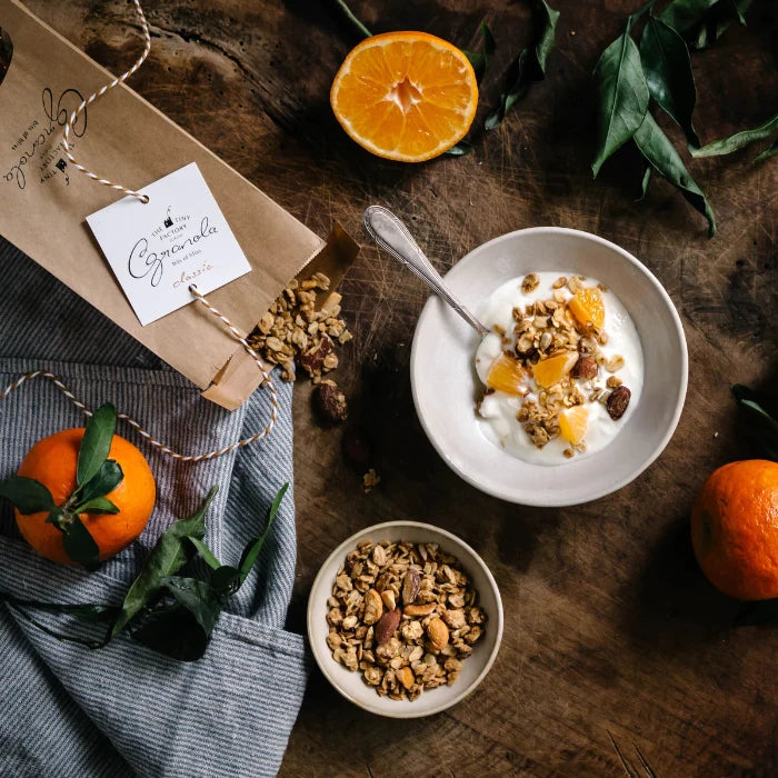 Ein Moodbild mit Classic Granola von der Tiny Factory, einer Schüssel mit Jogurt und Granola, Granola, Orangen auf einem holzigen Untergrund.