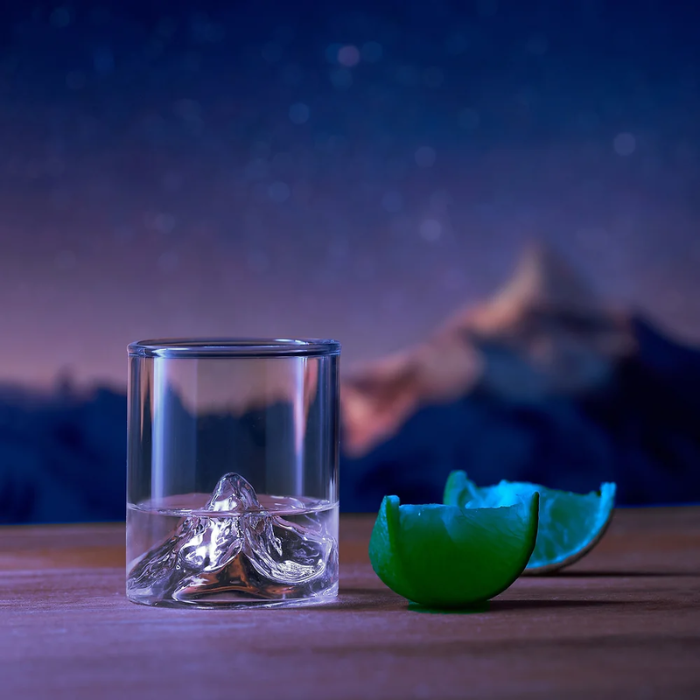 Schnappsglas Matterhorn befüllt mit Limette nebendran vor einem Sternenhimmel und dem Matterhorn im Hintergrund.