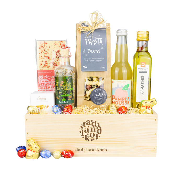 Bunte Osterglücksmomente Geschenkkorb vor weissem Hintergrund befüllt mit weisser Schokolade, Blüemli Pasta, Haldi Salz, Straumann Hüppen, Limonade, Rosmarinöl und Lindt Schokolade