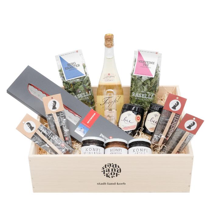 Family-Brunch Deluxe Geschenkkorb alkoholfrei mit Taucherli Heissi Schoggi, PanoramaKKnife, drei saisonalen Konfitüren, Bio Raselli Bergkräutertee, zwei Honig von der Bee Family, Bio Raselli Verveine Tee und alkoholfreier Schaumwein Tröpfel der ERSTE in einem Stadt KLand korb Geschenkkorb aus Holz vor weissem Hintergrund